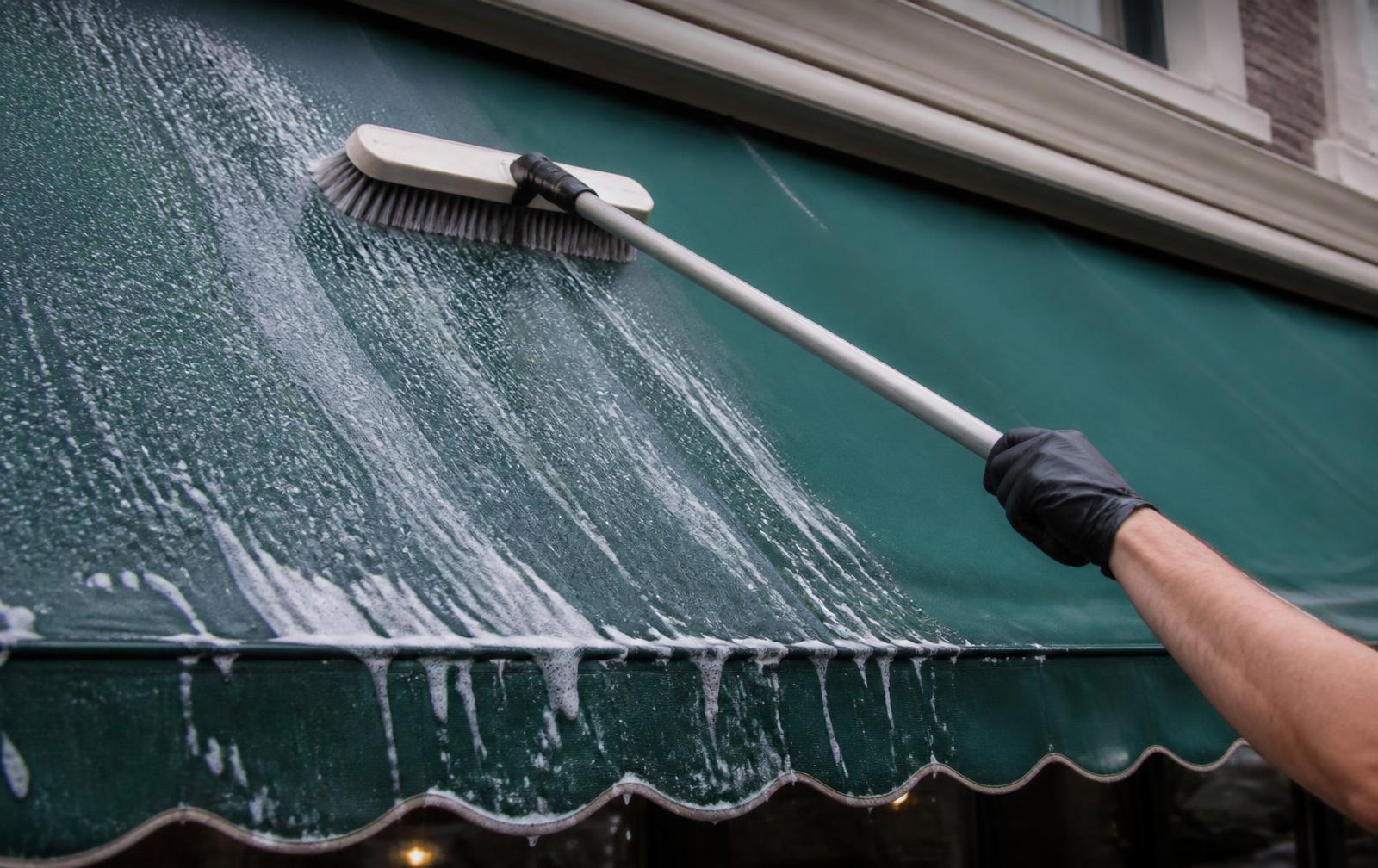 Technician scrubbing suds across a green fabric storefront awning with a long-handled soft brush